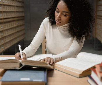 A person sits in an archive and writes in a book whilst sitting at a wood table. Some open, some closed and some stacked books lay on top of the table.