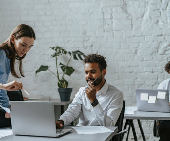 Two people sit behind desks working on laptops. One person sits on top of a desk on the left, pointing at a laptop screen.