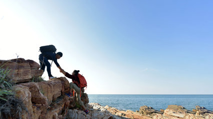 Two hikers on a seaside cliff on a sunny day. One is assisting the other by taking their hand and pulling them up.
