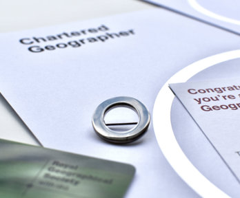 A silver circle lapel pin, a symbol of Chartered Geographer status, next to a card that says 'Congratulations, you're a Chartered Geographer'.