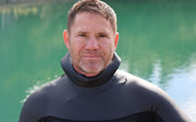 Steve Backshall facing the camera standing next to a body of water.