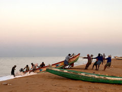Two fishing boats on a beach pictured with people on either side pulling it out of the water.