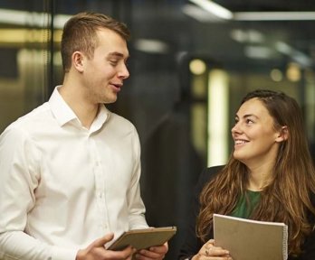 Two people in office setting in lively conversation while holding notebooks.