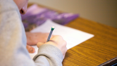 A student writes on an exam paper whilst being sat at a wooden desk.