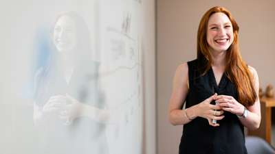 A teacher stands next to a blurred out white board whilst holding a white board marker.