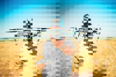 A man standing in yellow field of wheat, holding a silver drone out towards the camera. The drone obscures his face.