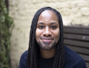Personwearing a dark blue shirt sitting on a wooden bench outdoors against a light stone wall.