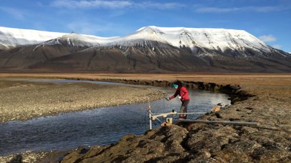 A researcher collecting water temperature measurements at the edge of a river in a baron landscape, with mountains in the background.