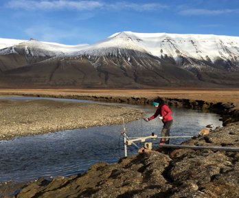 A researcher collecting water temperature measurements at the edge of a river in a baron landscape, with mountains in the background.
