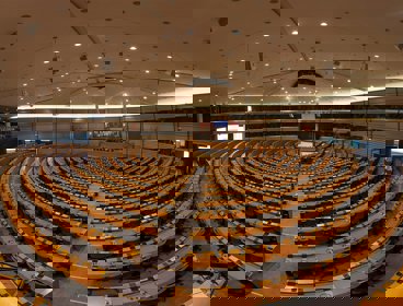Circular rows of seats in a conference room with a stage at the front. There are European flags behind the stage.