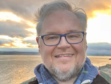 Person wearing a green jacket and blue scarf standing by a lake at sunset.