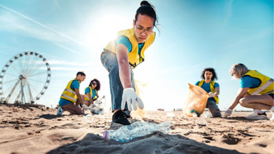 People picking up litter on a beach on a sunny day. In focus is a person reaching for a plastic bottle.