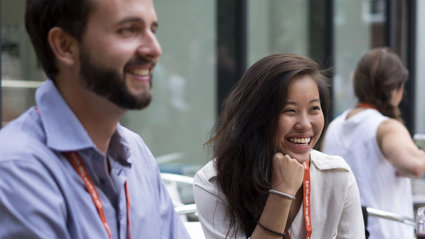 Two people sit next to each other smiling while wearing professional clothing and lanyards.