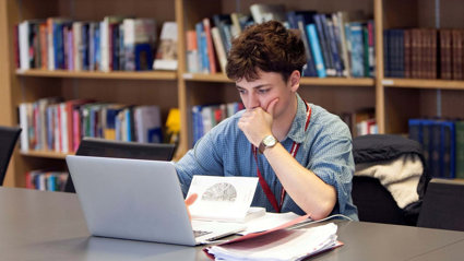 Person sat at a table looking at a laptop screen with notepads and books surrounding the laptop. Behind the table there are bookshelves with stacks of books.