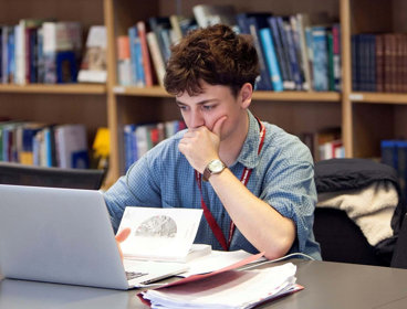 Person sat at a table in a library looking at a laptop screen with notepads and books surrounding the laptop.