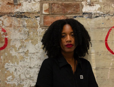 Person wearing a black polo shirt standing against a brick wall with red paint.