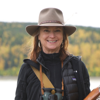 Headshot of Roseann Hanson. She's in the outdoors, wearing a fedora and binoculars.