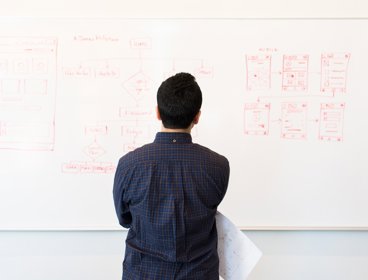 Man holding a piece of paper looking at a whiteboard with diagrams drawn on it.