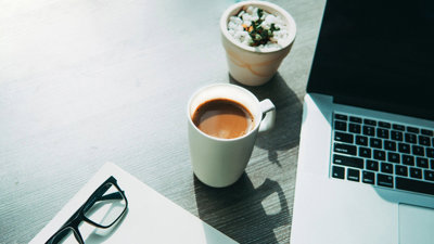 A cup of coffee, glasses, a sheet of paper, a cactus and a laptop on top of a wooden desk.