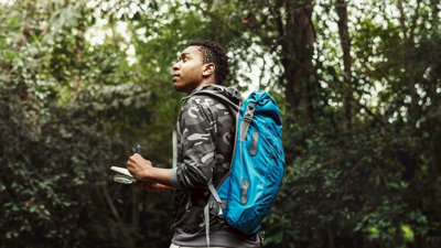 Person with a backpack on in a forest writing something down in a notebook while looking up.