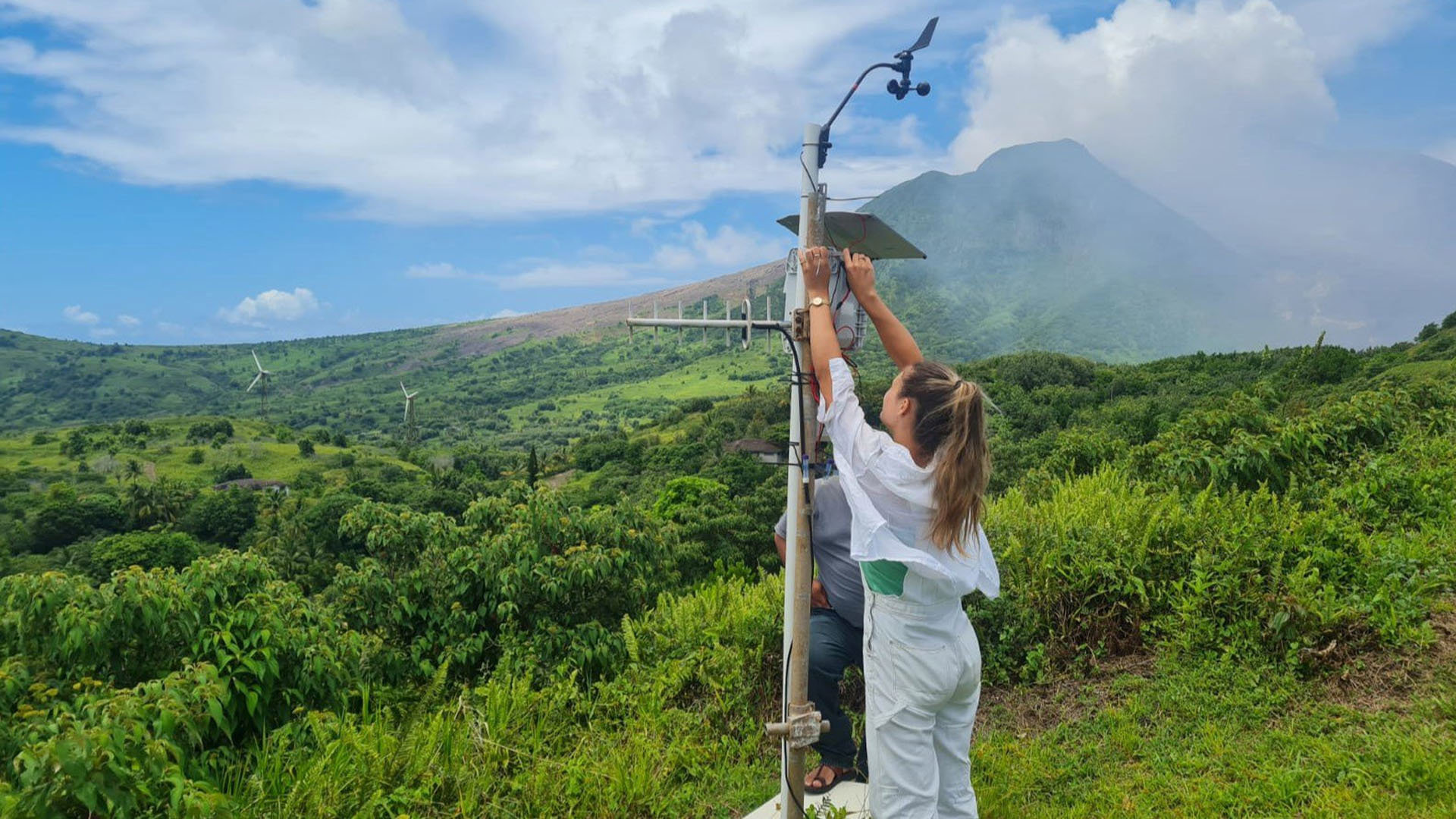 Two people with an air quality monitoring device in St George's Hill, Montserrat. Volcanic plume rises in the background.
