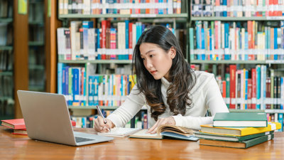 Student in a library looking at a laptop screen while writing notes with in the background various books on a book shelf.