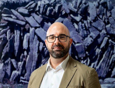 Person in beige blazer and white shirt standing in front of a large abstract painting with dark, jagged shapes.