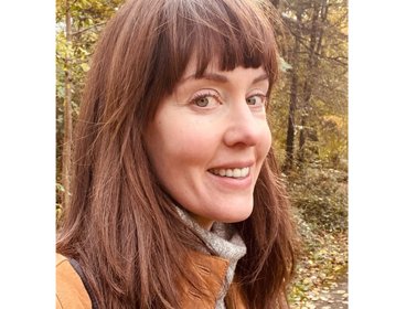 Person wearing a tan jacket and gray scarf standing on a forest path covered with autumn leaves.