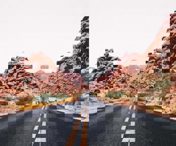 A Nevada road stretching out into the distance, the yellow central lines weaving slightly where the road bends. The road has rocky outcrops either side and in the distance