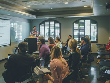 A group of people sat in a room with a projector screen with one person in the front presenting.