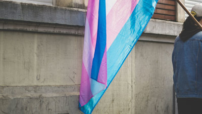 A blue, pink, and white trans flag hangs via a flag pole is held by a person faced away from the viewer and partially cut out of frame.