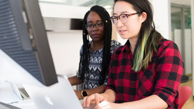 Two people sit next to each other at a desk. one uses the computer whist the other watches them.