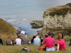 Young people sitting on a grassy sloped hill and listen to a teacher talking about the natural landscape