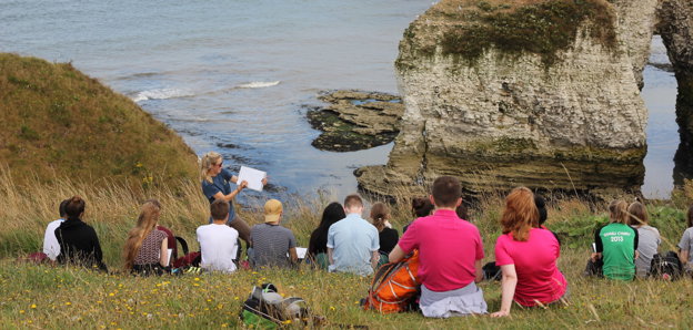 Young people sitting on a grassy sloped hill and listen to a teacher talking about the natural landscape