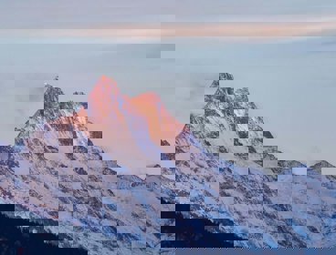 A mountain in the early or late sun, with its tip in soft sunlight. The mountain is purple, blue and white in colour where the rocks and snow meet.