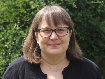Person wearing a black top standing outdoors with green foliage in the background.