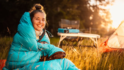 A person in a sleeping bag sitting in the outdoors next to a tent and foldable picknick table on a sunny day.