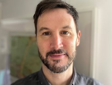 Person wearing a dark button-up shirt standing indoors with a blurred background of a room.