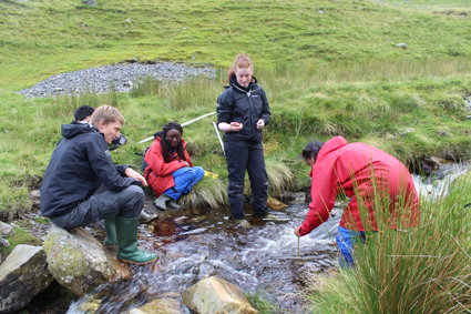 A group of students are gathered by a small stream, one is measuring the depth of a water using a stick. The water is fairly fast moving.