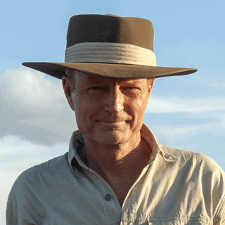Headshot of Jonathan Hanson on a sunny day in the outdoors. He's wearing a brown fedora and a safari style blouse.