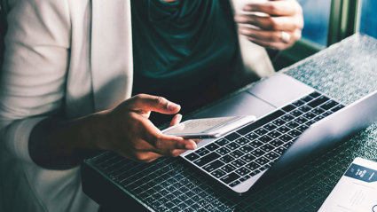 A close-up of a person holding a phone while sat at a desk with a laptop.