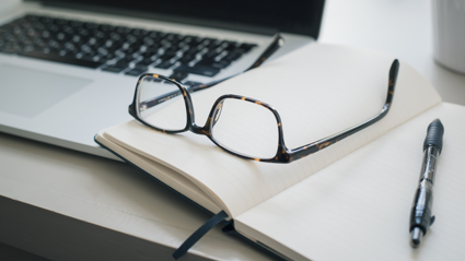 A pair of glasses, open laptop, empty notebook and a pen messily lay on top of each other on a plain grey surface.
