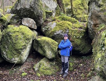 Person in blue jacket standing amongst large mossy rocks.