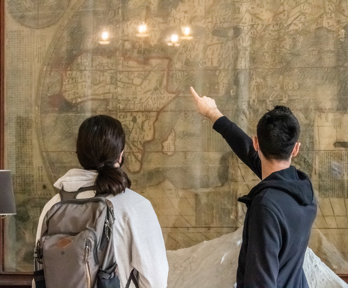 People stand in from of a large wall mounted map with their back to the camera as they look and point at the map.
