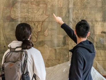 People stand in from of a large wall mounted map with their back to the camera as they look and point at the map.
