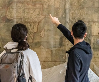 People stand in from of a large wall mounted map with their back to the camera as they look and point at the map.