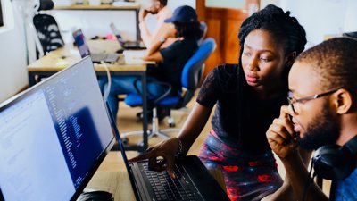 A Black man and woman sat looking at a laptop in an office setting.