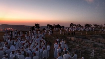 A large group of people gathered for a ceremony on a mountaintop as the sun is setting.