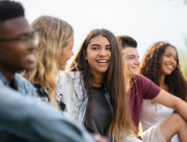 Group of five young people sitting outdoorsk, with the focus is on a brown-haired person in the centre.