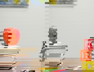 A wooden school desk with an apple placed on top of a pile of books. Some coloured pencils are also seen, as well as building blocks with A, B and C on them, placed in a stack